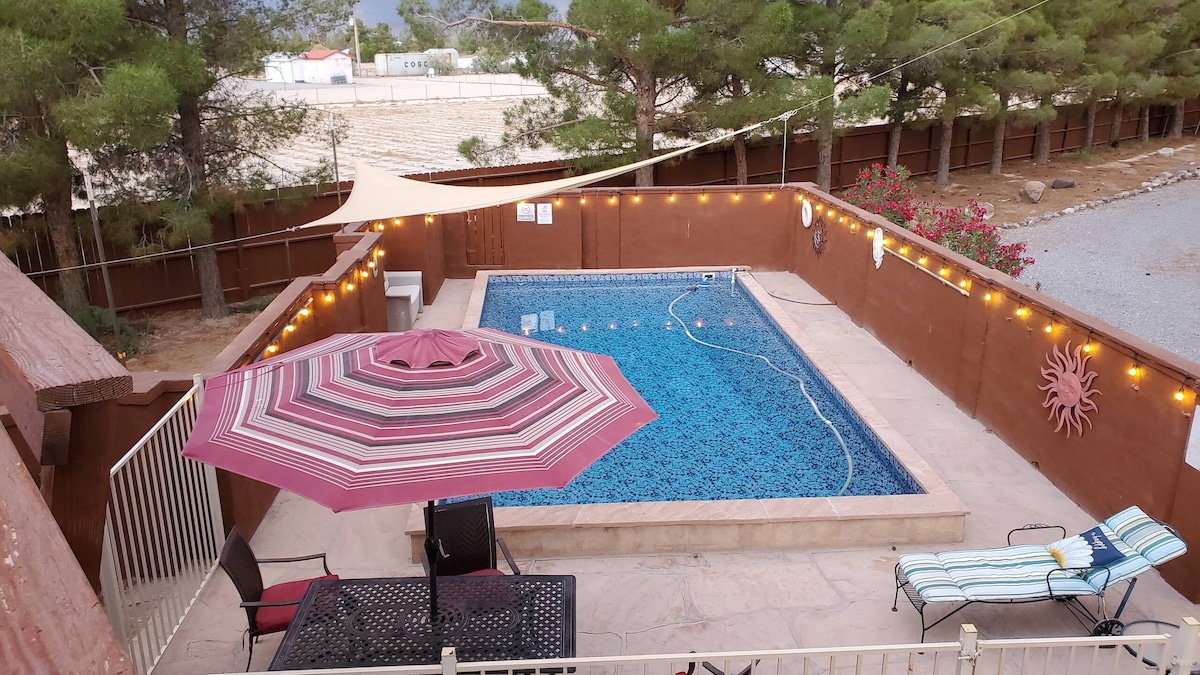 A private pool is surrounded by a spacious patio lit with string lights, creating a welcoming outdoor space. A pink and white striped umbrella provides shade over a table and chairs, while a lounge chair rests nearby for relaxation. Desert landscaping and tall trees frame the area.