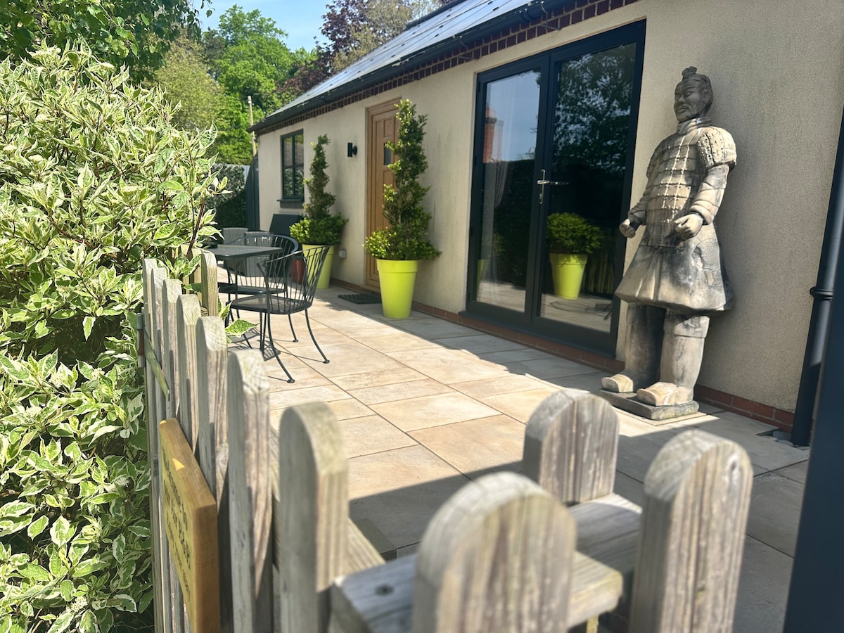 A patio area is visible, featuring a paved surface and a small table with two chairs. Potted plants are placed near the entrance, while a decorative statue stands prominently beside the door. A wooden fence frames the scene, complementing the lush greenery surrounding the space.