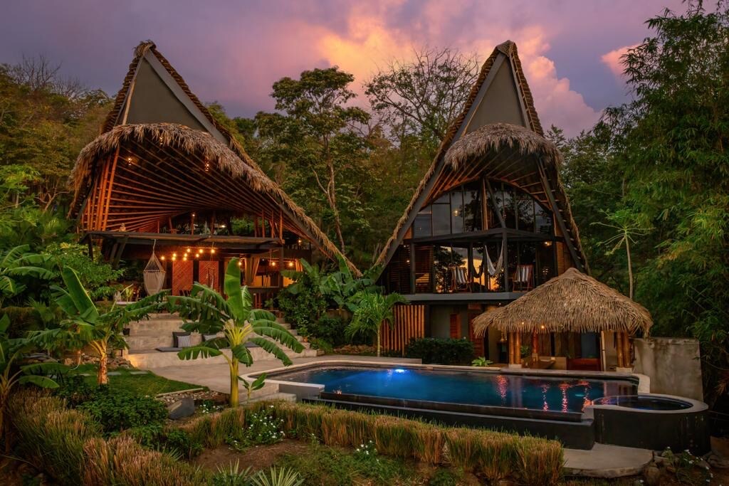 The image captures unique eco-tropical cabañas with straw roofs, blending harmoniously with lush greenery. A saline infinity pool is located in the foreground, surrounded by well-maintained gardens and tropical plants. The sky showcases a gradient of colors as the sun sets behind the structures.