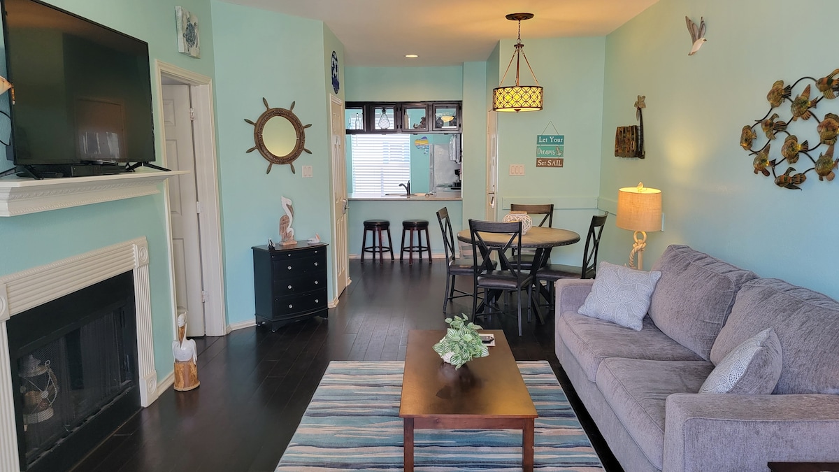 An open living area features a cozy gray sofa, a coffee table on a blue-striped rug, and a stylish dining set. The room is illuminated by a pendant light, with a flat-screen TV mounted above a fireplace. A kitchen is visible in the background.