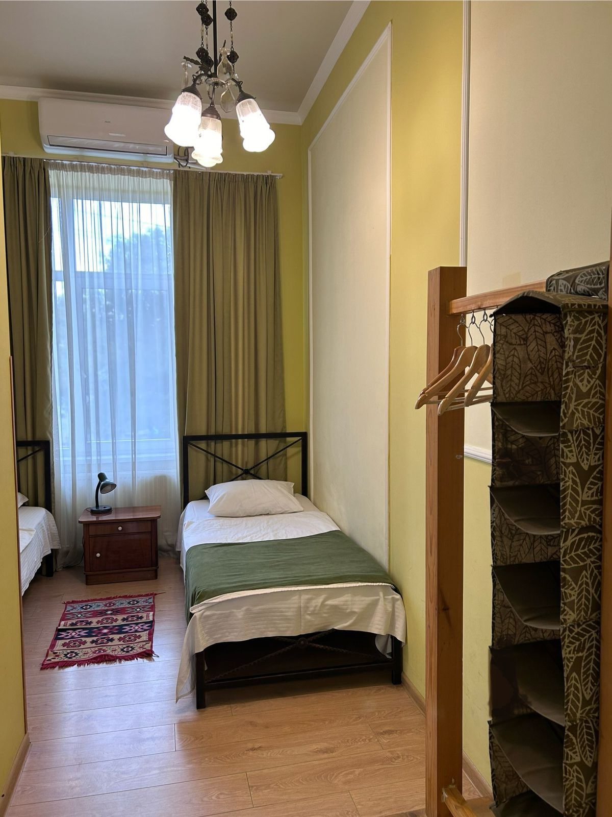 A bright room features two single beds arranged side by side, each topped with white linens and a green throw. A wooden nightstand holds a small lamp, and light flows in through the tall window adorned with soft curtains. A hanging rack displays garments on the right.