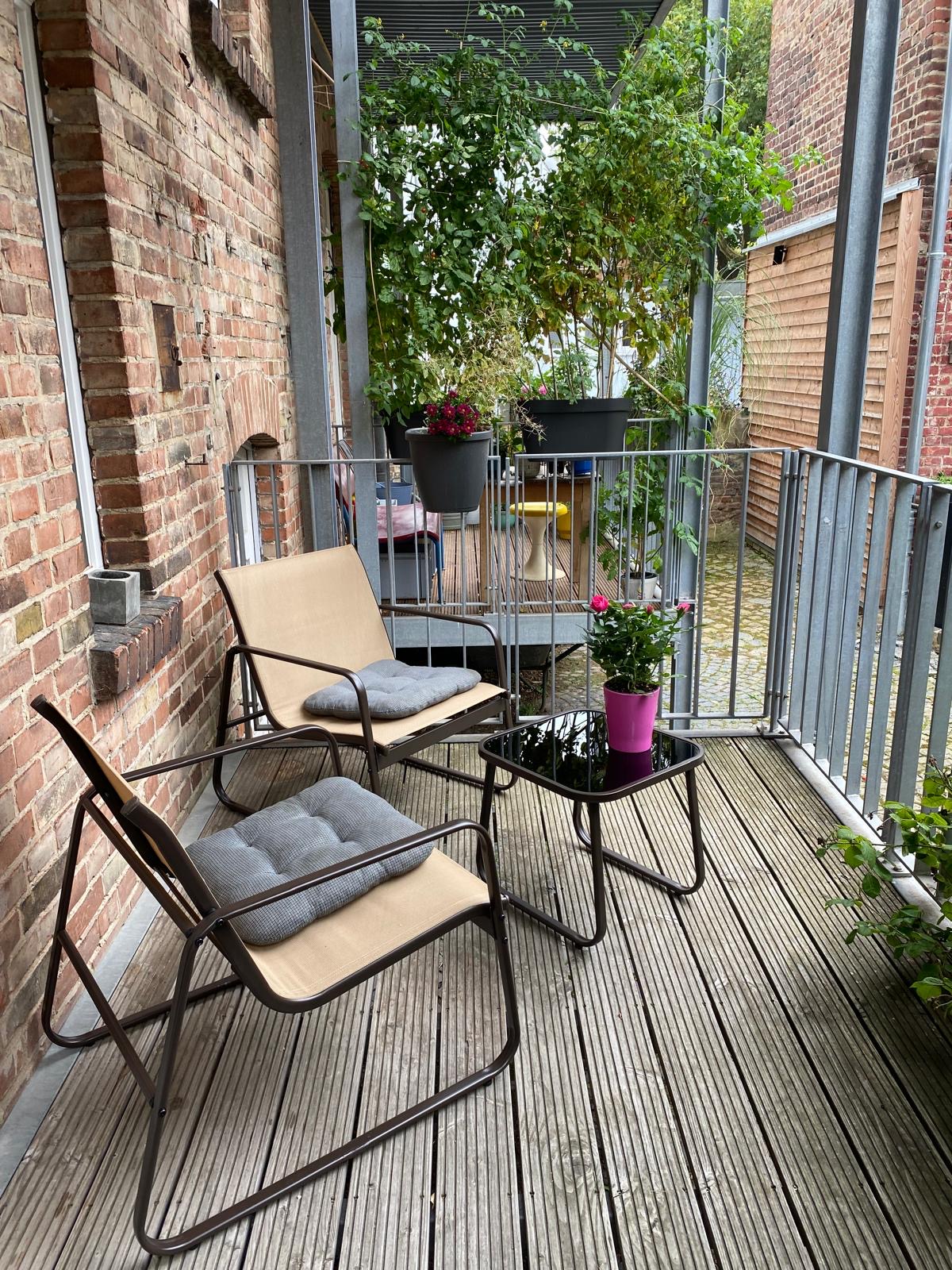 A balcony area is shown, featuring two folding chairs and a small glass table. Potted plants add greenery, while wooden decking creates a warm setting. The surrounding walls are made of exposed brick, contributing to a rustic feel.