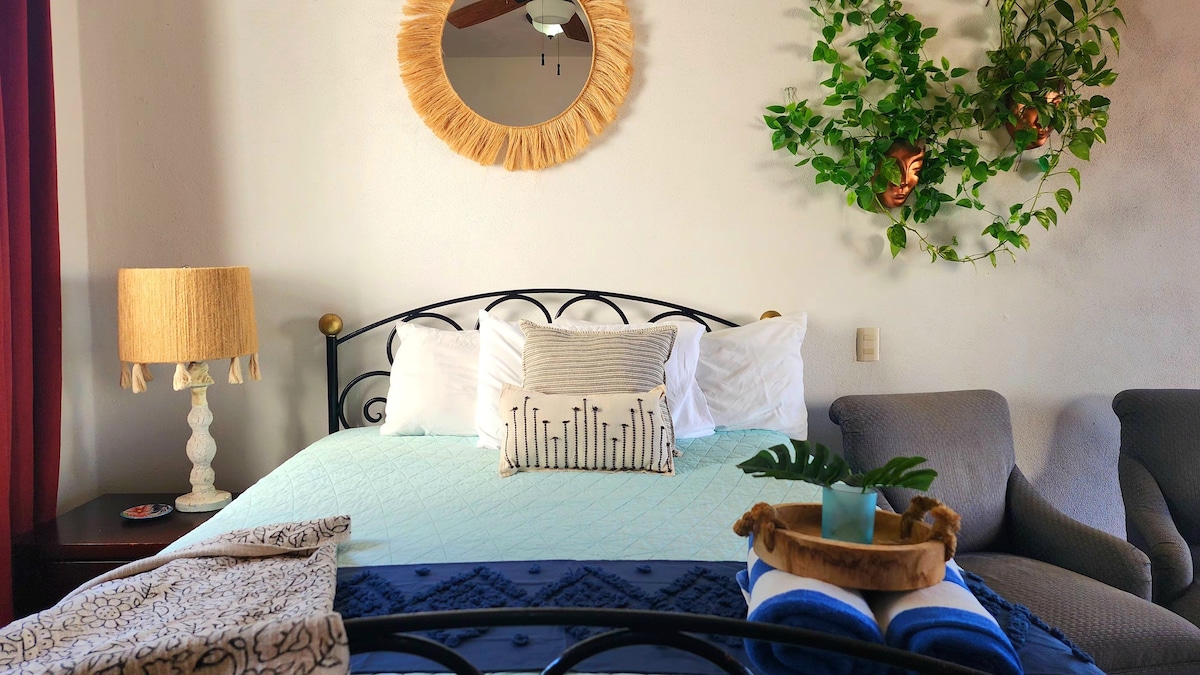A cozy studio apartment features a wrought-iron bed covered with a light blue quilt. Decorative cushions are arranged atop the bed. A lamp with a textured shade and a round mirror adorn the space. Greenery adds a fresh touch to the wall behind a comfortable armchair.