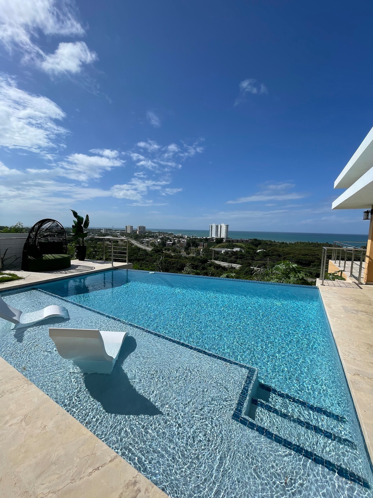 A private infinity pool is framed by a spacious patio, showcasing clear blue water that reflects the sky. Surrounding greenery adds a natural touch, while modern lounge chairs invite relaxation. The landscape extends into distant city views and the ocean, enhancing the tranquil setting.