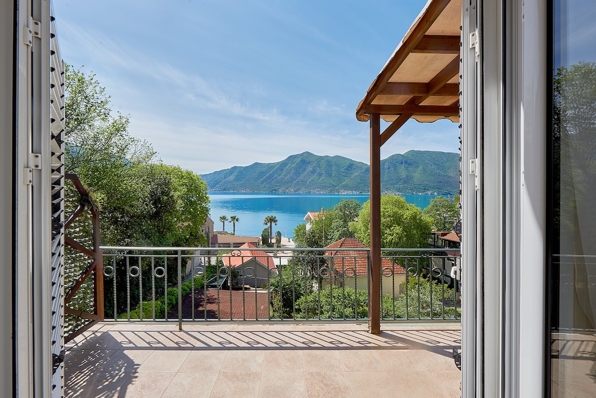 A balcony view captures the serene landscape featuring lush green mountains and a sparkling blue sea. The scene includes glimpses of terracotta rooftops beneath the expansive sky, emphasizing the tranquil setting of the coastal village.