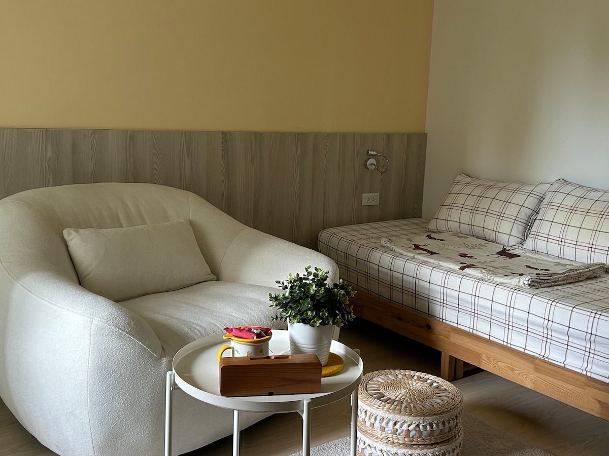 The room features a soft, oversized chair and a wooden bed, dressed in checkered bedding. A small round table holds a decorative potted plant and a tray. A woven storage basket complements the space, creating a cozy and functional environment.