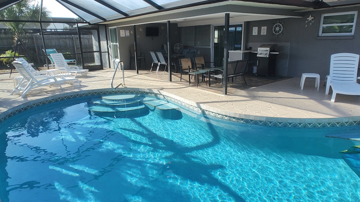 An inviting screened-in pool area features a large pool with steps and a shallow seating space. Surrounding it, lounge chairs await sunbathing, and a covered patio offers seating and a grill for outdoor dining. Lush landscaping adds a tropical touch.