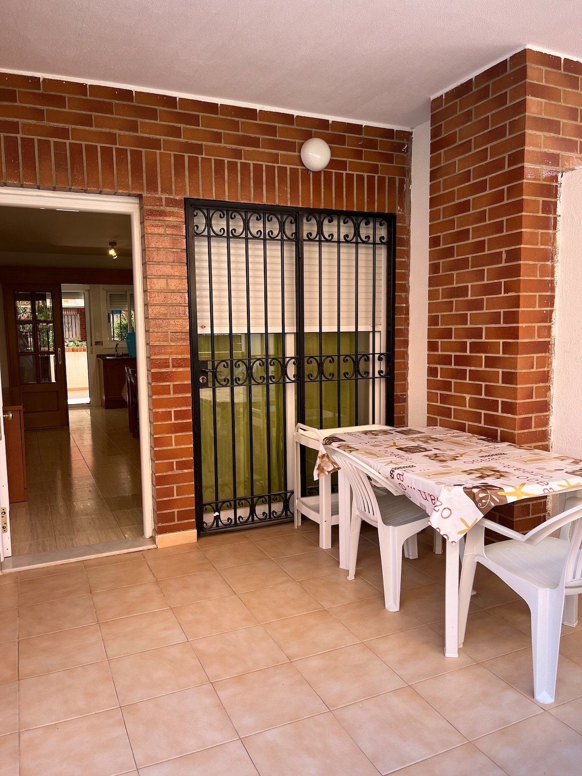 A covered terrace is presented, featuring a table surrounded by four plastic chairs. The floor is tiled, and the walls are adorned with red brick. A security gate leads to the interior of the apartment, providing a blend of indoor and outdoor space.
