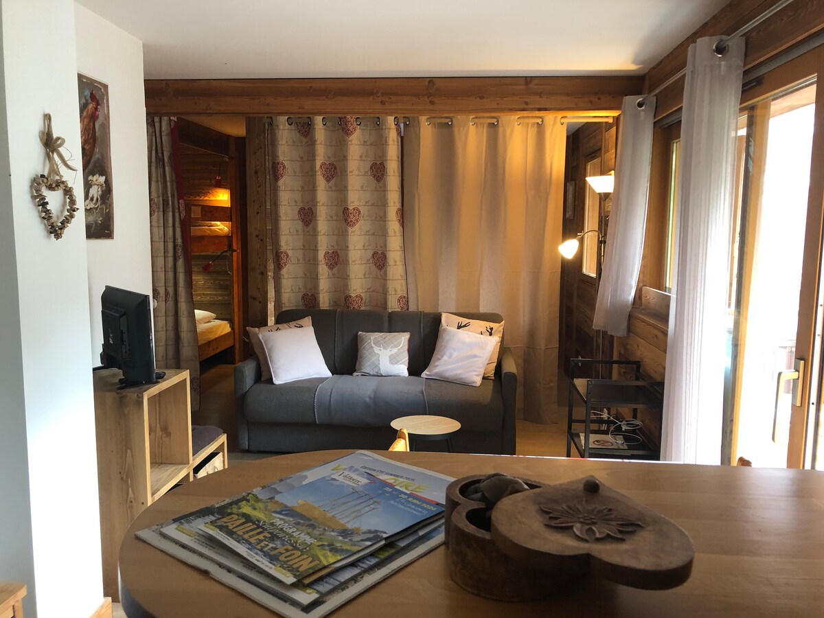 A cozy living area is featured, showcasing a gray sofa complemented by two white cushions. Light floods in through large windows adorned with sheer curtains. A small round table sits nearby, with magazines arranged on a wooden surface, while a warm, inviting ambiance is created by soft lighting.