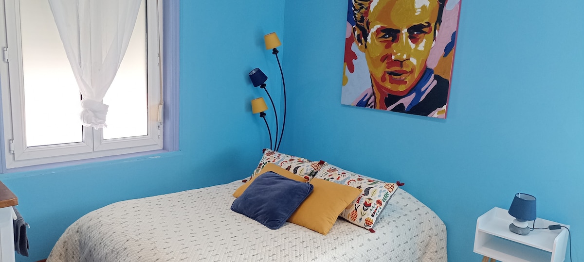 A cozy bedroom features blue walls and a colorful artwork above the bed. A spacious bed is adorned with patterned pillows in various colors. Natural light enters through a window, which is dressed with sheer curtains, complementing the simple bedside table and lamp.