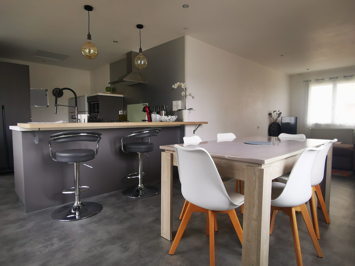 An open kitchen and living area is presented, featuring a sleek countertop with high stools and a wooden dining table surrounded by white chairs. Modern appliances and decor are visible, with natural light entering through windows, creating a bright and functional space.