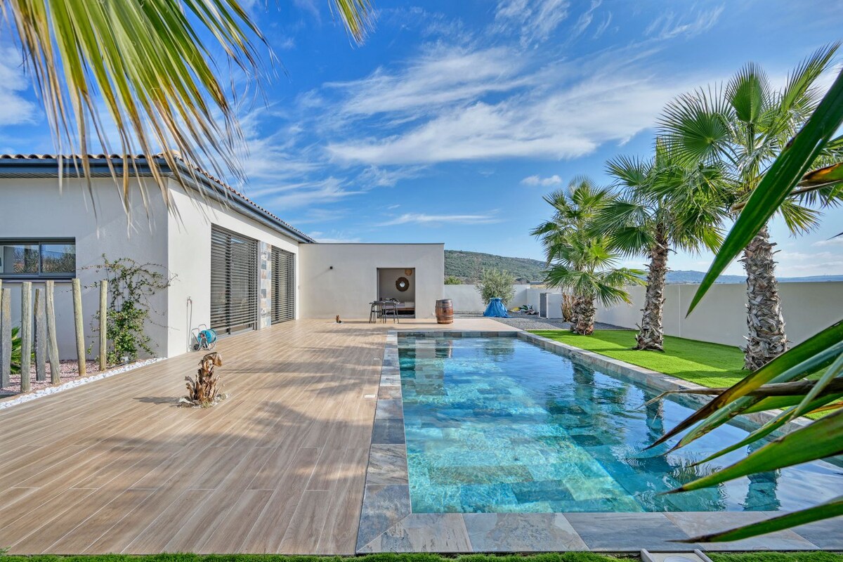 An outdoor space features a sleek swimming pool surrounded by a wooden deck and lush palm trees. The clear blue water reflects the sky, while a spacious patio area offers potential for relaxation and outdoor dining.