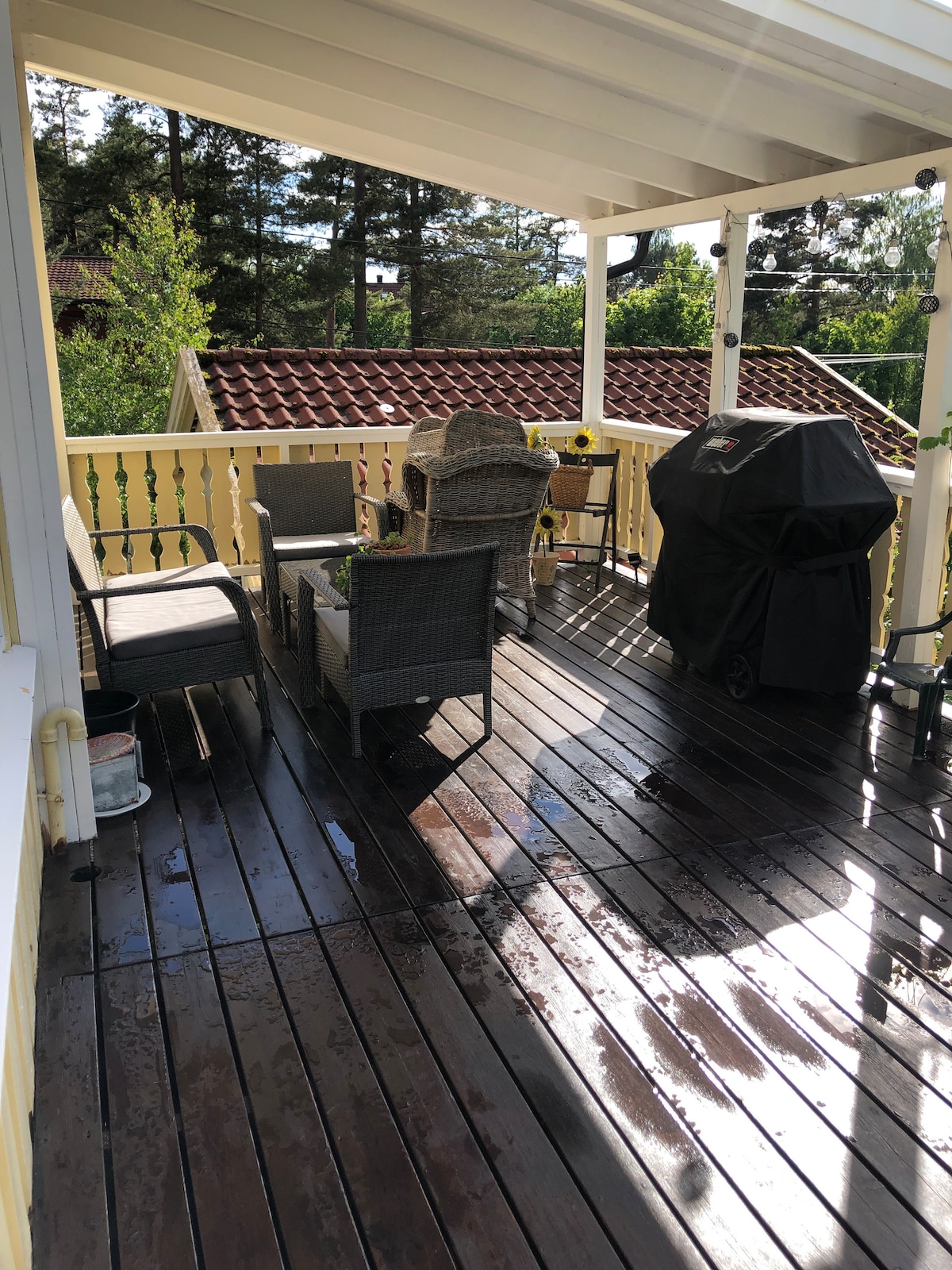 A spacious outdoor area features a wooden deck with seating arrangements including woven chairs and a table. A grill is positioned nearby, set against a backdrop of greenery and rooftops. Sunlight reflects off the polished surface, creating a bright and inviting space.