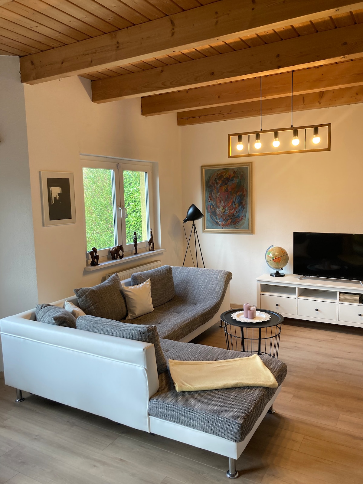 The living area features a sectional sofa with light tones and plush cushions, arranged to encourage relaxation. A modern floor lamp adds warmth, while a television is mounted on a white media console. Natural light enters through a window, framed by a view of greenery.