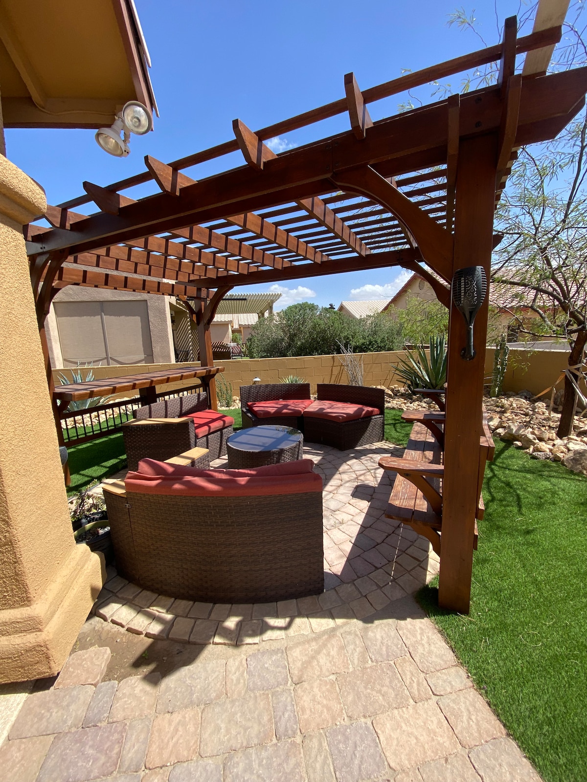 A shaded seating area features a wooden pergola supported by beams, providing shelter over a circular arrangement of dark wicker chairs and a central table. Surrounding elements include green grass and desert landscaping with rocks and sparse plants, creating a private outdoor retreat.