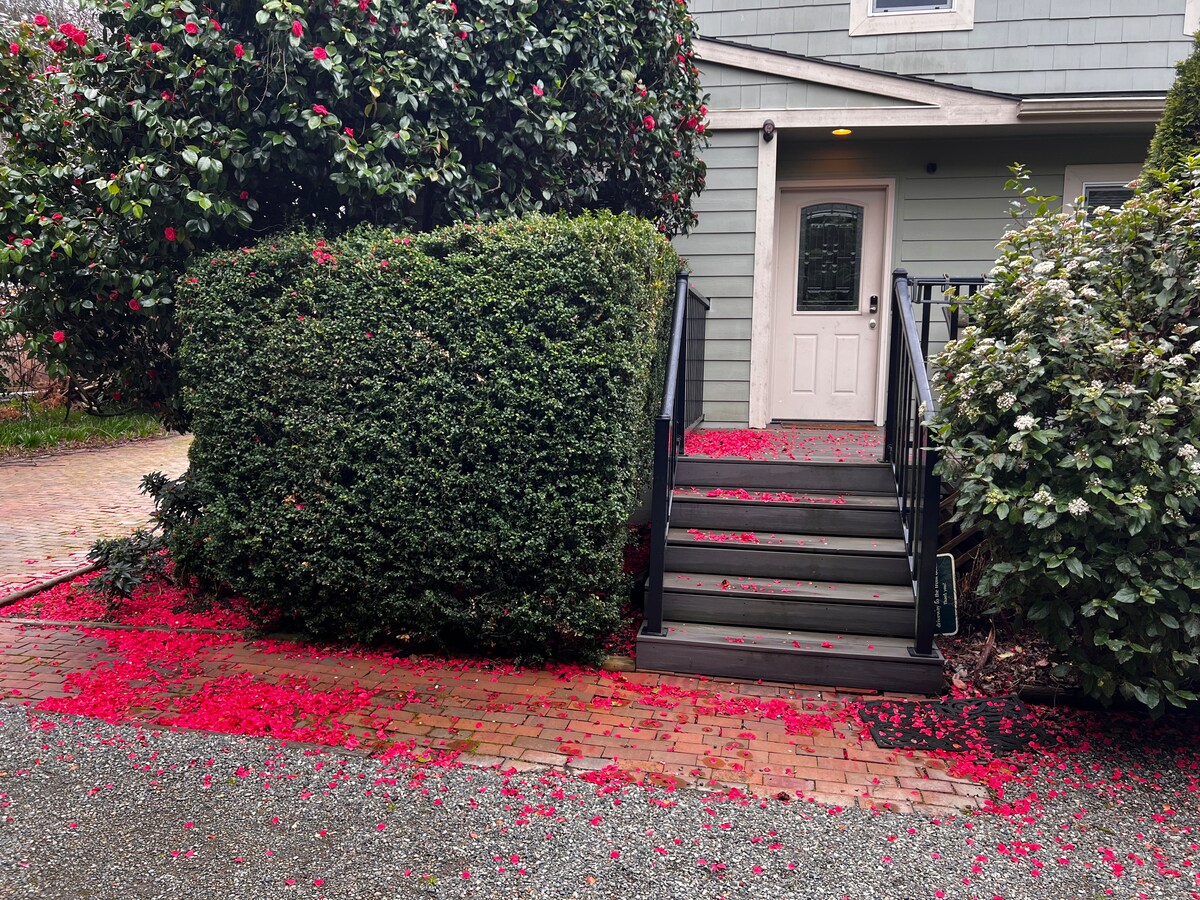 A winding pathway leads up to a home entrance, framed by lush greenery and flowering shrubs. Bright red petals cover the steps and ground, creating a vibrant contrast against the gray brick and gravel surfaces, inviting guests into the space.