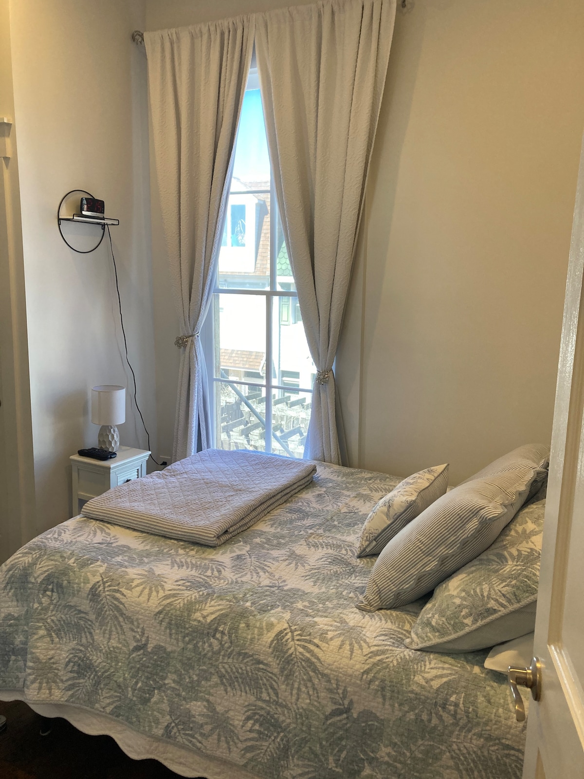A cozy sleeping area features a spacious bed dressed in soft, patterned linens. Light-colored curtains frame a window that invites natural light, while a bedside table holds a lamp. A folded blanket is neatly positioned on the bed, adding to the welcoming ambiance.