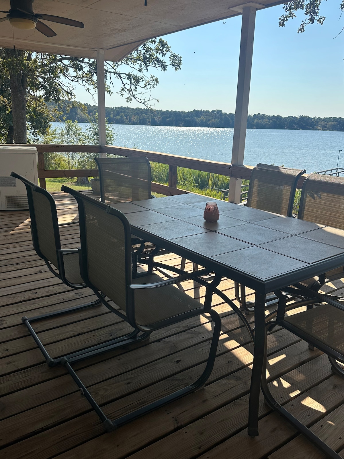 A spacious patio deck is highlighted, featuring a large dining table surrounded by several chairs. The deck offers a view of the tranquil waterfront, with lush greenery bordering the area. Natural light reflects off the water, creating a serene environment for relaxation.