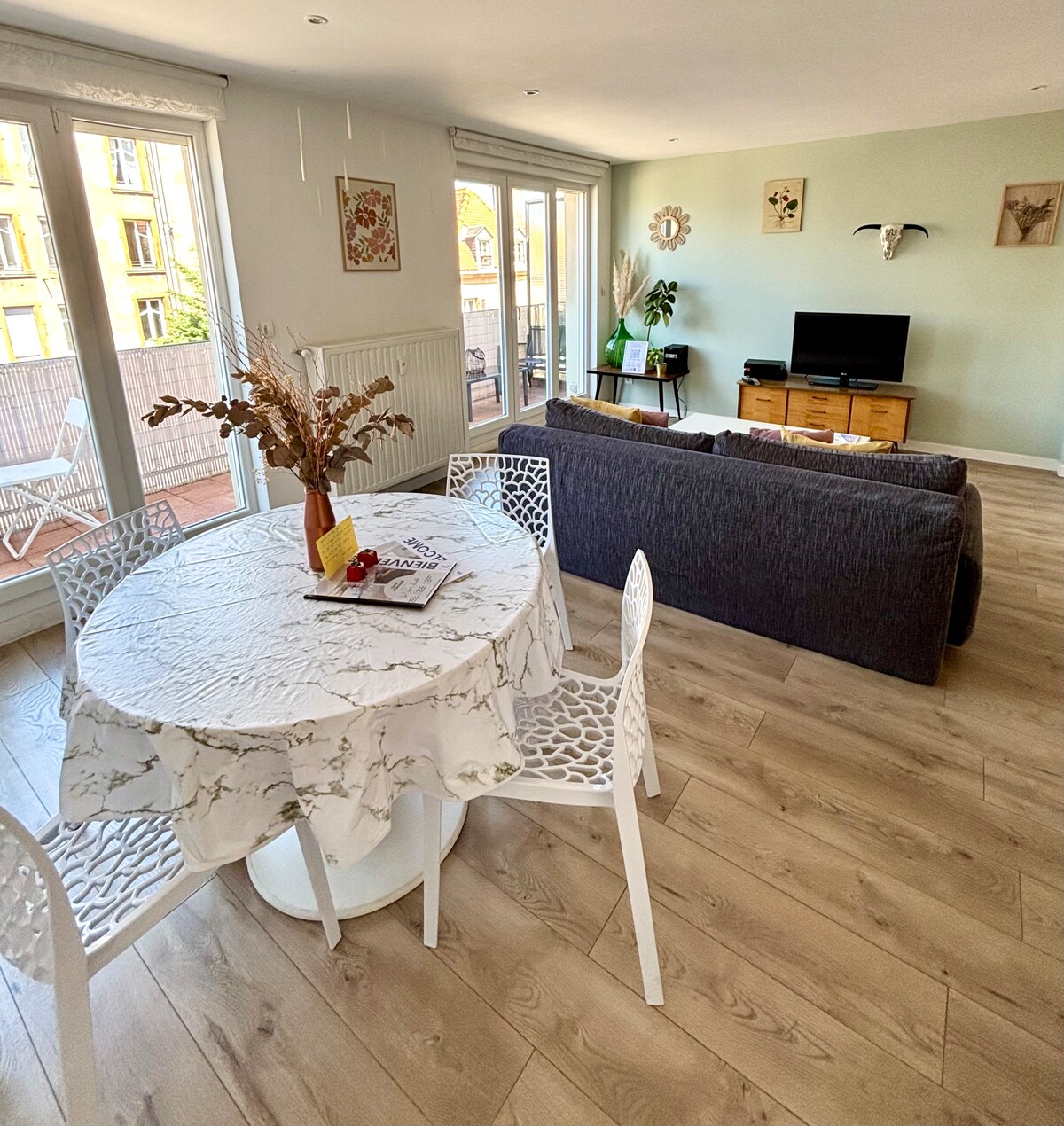 A modern living area features a round dining table with a marble top and four white chairs, complemented by a dark sofa. Large glass doors provide access to a balcony, allowing natural light to fill the space. A television is positioned on a wooden console.