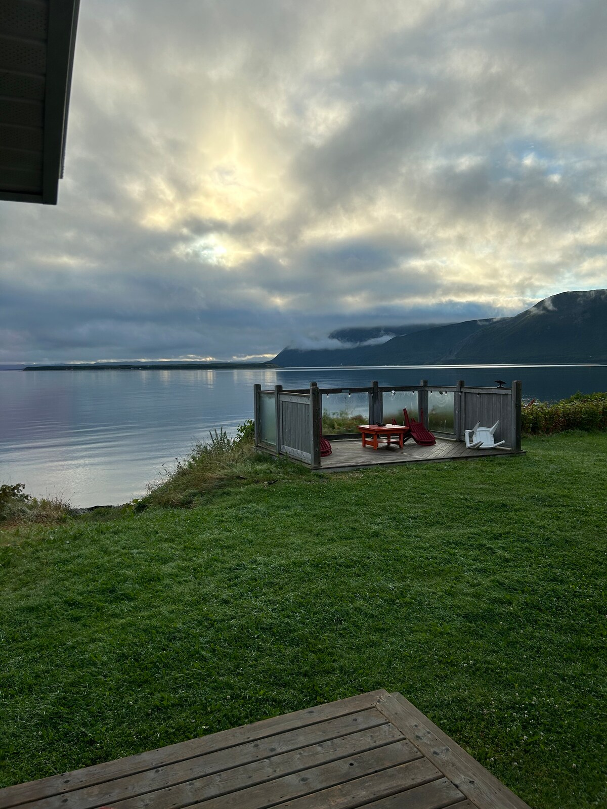 An outdoor patio area is visible, featuring a wooden deck and two red chairs. A large grassy expanse leads to the bay, where calm waters reflect the soft hues of the cloudy sky. Mountains can be seen in the distance, adding to the serene view.