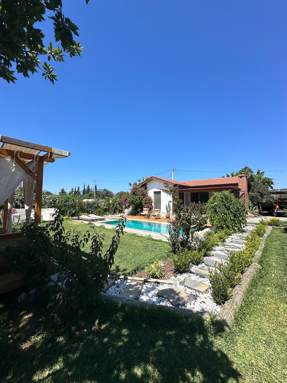A charming residential property is surrounded by lush greenery and features a serene pool. A wooden structure offers shade, while a manicured garden with stone pathways leads to the house. Clear blue skies enhance the tranquil atmosphere of this inviting outdoor space.