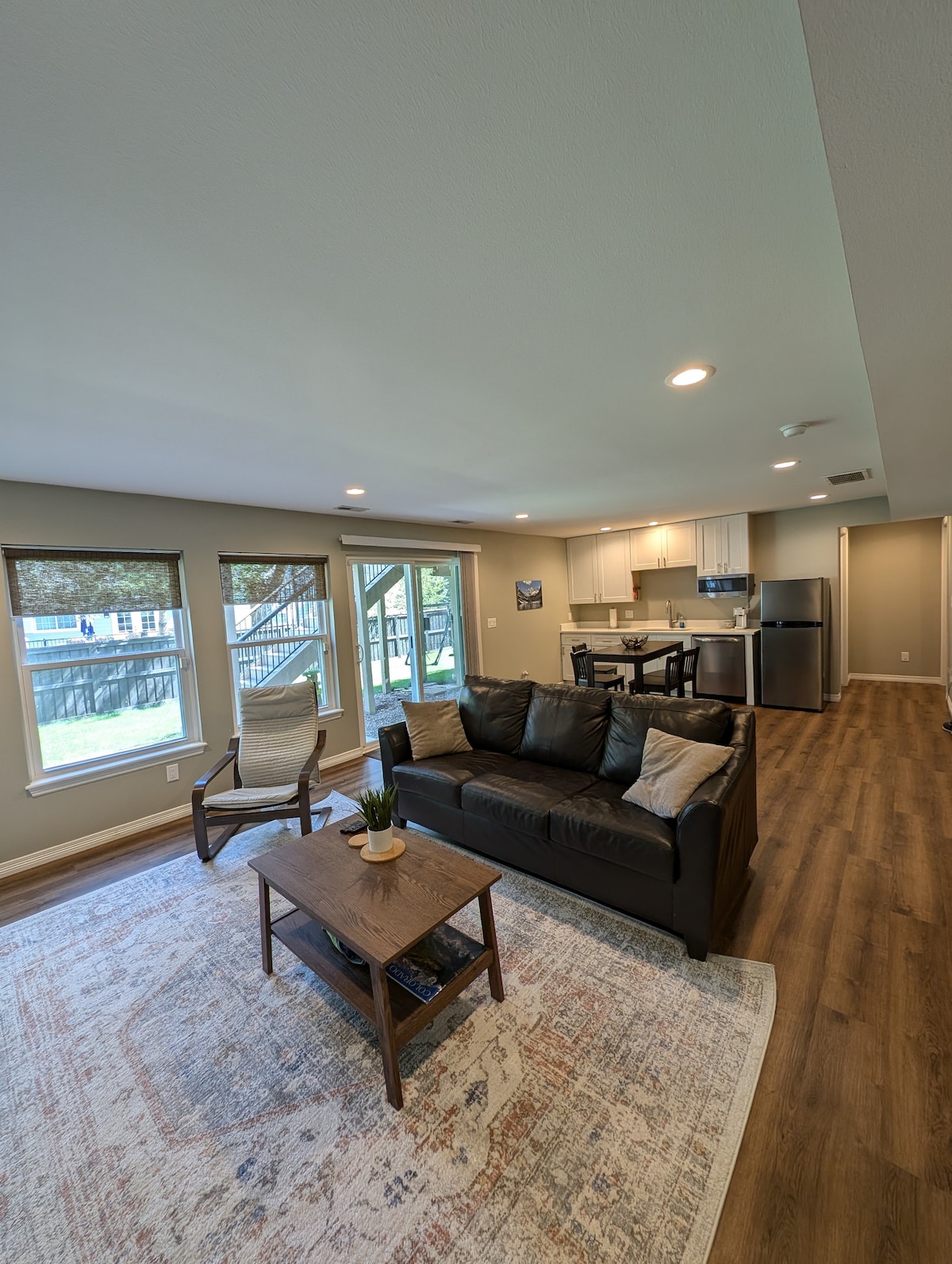 A spacious living area features a dark sofa and a wooden coffee table, complemented by an area rug. Large windows allow natural light to illuminate the space, which leads to a kitchen area with modern appliances and dining seating in the background.