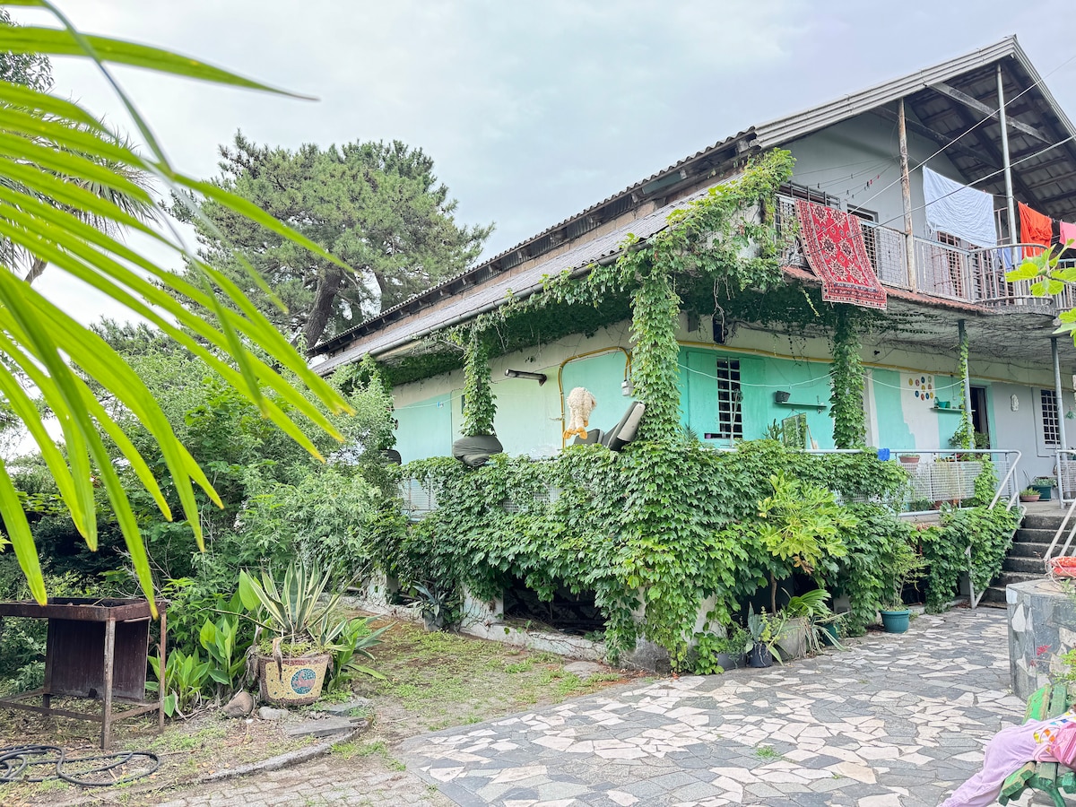 The house is surrounded by lush greenery, with vines climbing the walls. A spacious balcony with seating overlooks the garden. Colorful textiles are hung out to dry, adding a relaxed vibe, while stone pathways lead through the vibrant outdoor space.