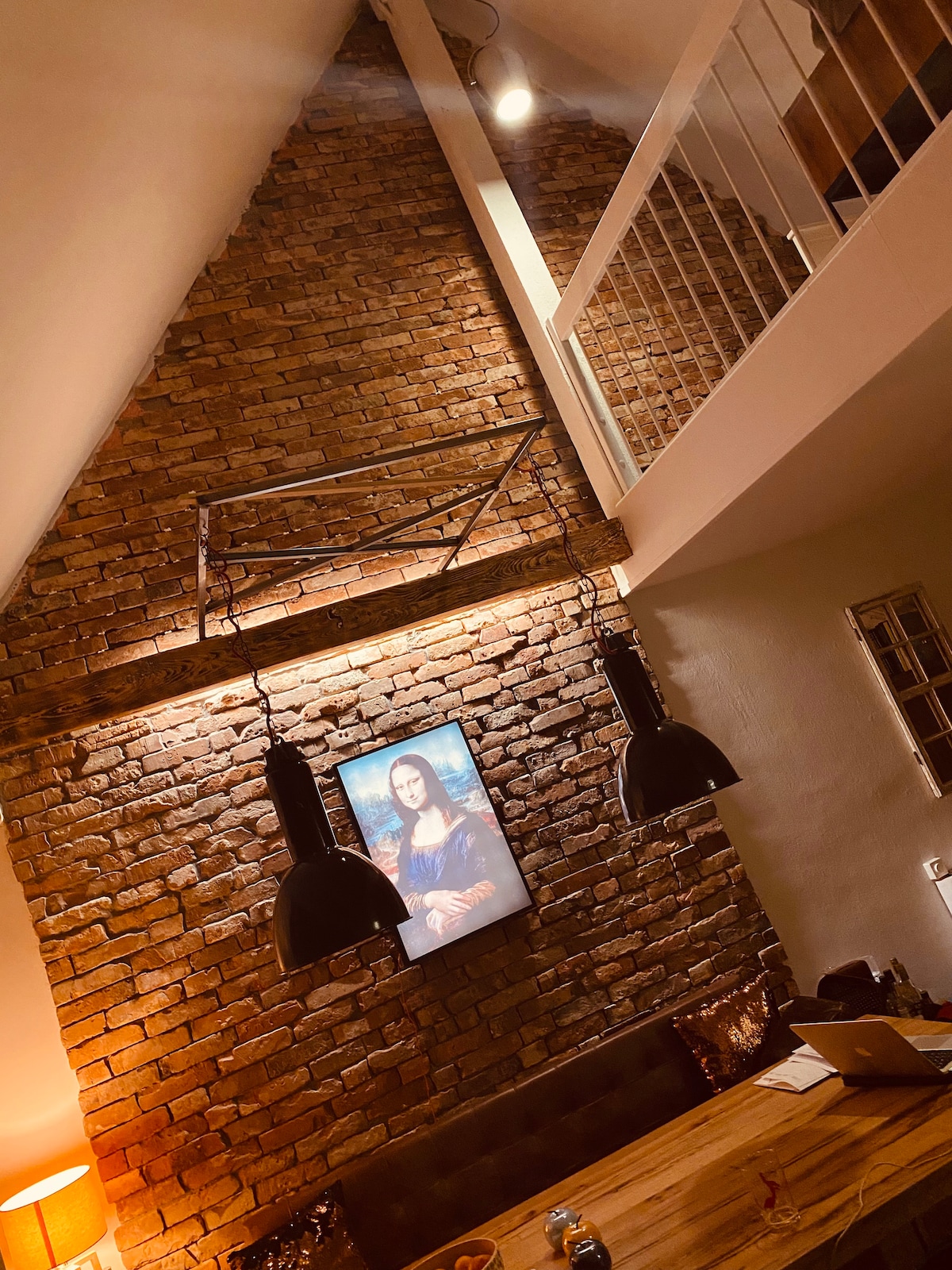The interior features a striking triangular brick wall, highlighted by warm lighting. Two industrial-style black pendant lamps hang above a wooden table. A framed artwork adorns the wall, contributing to the overall design of the space.