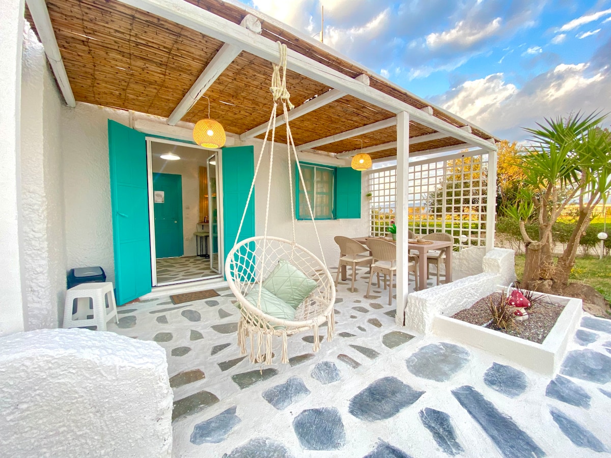 A charming outdoor patio features a hanging chair and a dining table surrounded by stone pavements. Bright turquoise doors and soft yellow pendant lights offer a welcoming atmosphere. Lush greenery and a small flower bed enhance the serene outdoor space, ideal for relaxation.