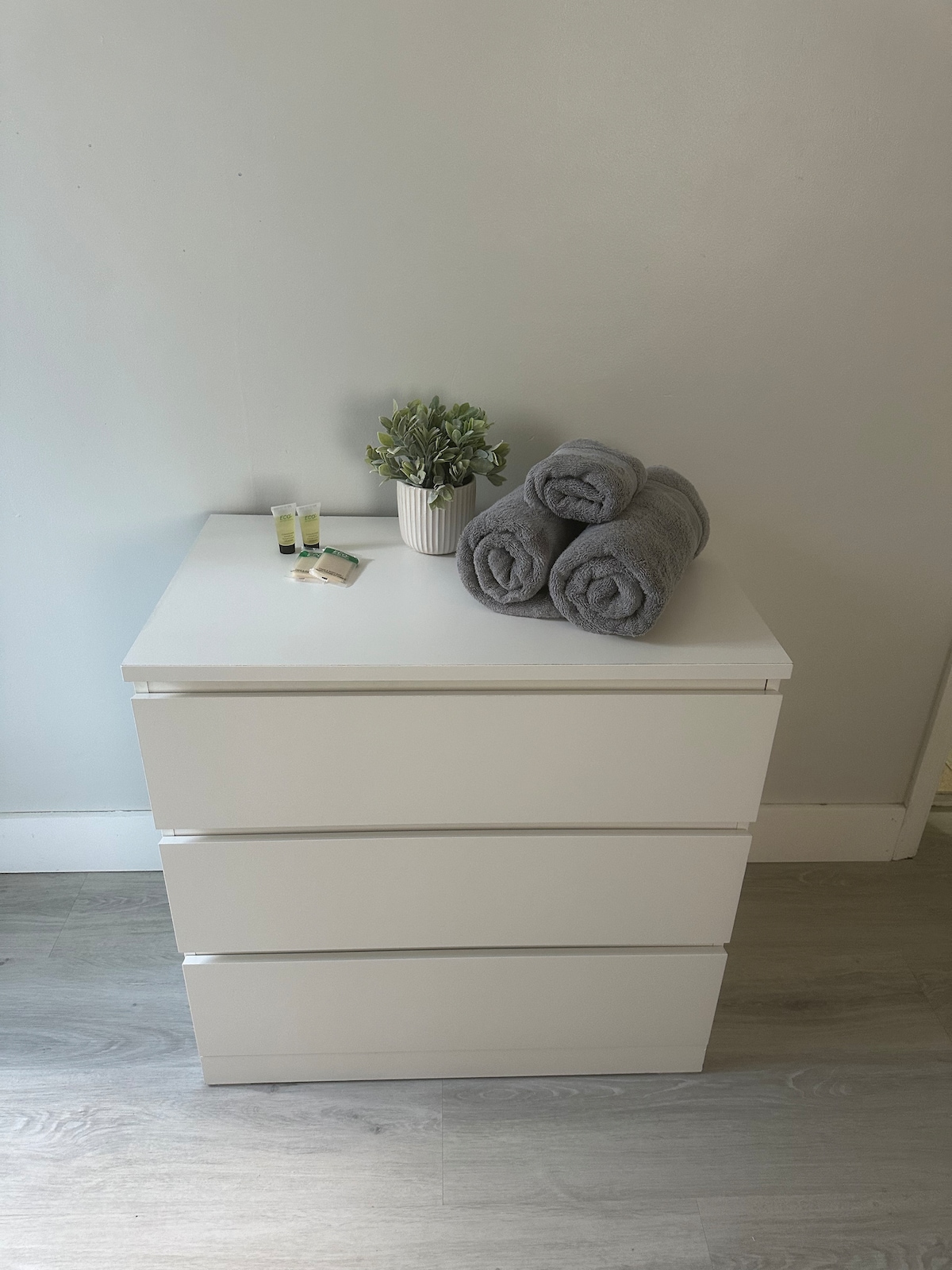 A minimalist dresser is positioned against a light wall. On top, neatly rolled gray towels are arranged beside a small potted plant and two travel-sized toiletries. The flooring is light wood, contributing to a clean and organized appearance.