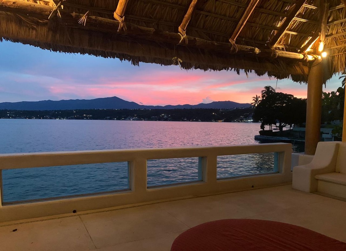 A covered outdoor seating area features a thatched roof, providing shade and comfort. A panoramic view of the water and distant mountains is visible, with a vibrant sunset casting warm hues across the sky. Soft lighting enhances the serene ambiance.