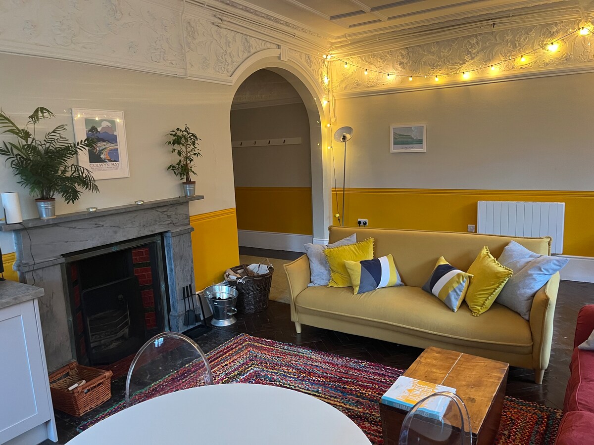 A vibrant living area features a yellow couch adorned with decorative pillows, providing a pop of color against neutral walls. A cozy rug lies on the polished floor, while soft lighting highlights the intricate ceiling details. A round table and modern lamp complete the inviting space.