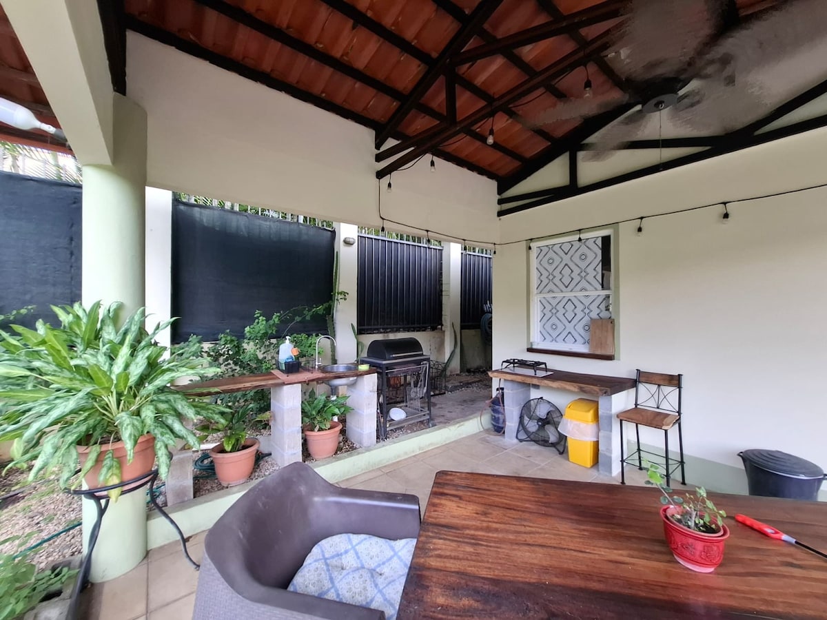 An open outdoor kitchen area features a wooden table and chairs, surrounded by potted plants. A barbecue grill is positioned nearby, with preparatory surfaces and a casual seating arrangement. The area is sheltered by a roof, providing an inviting space for meals.