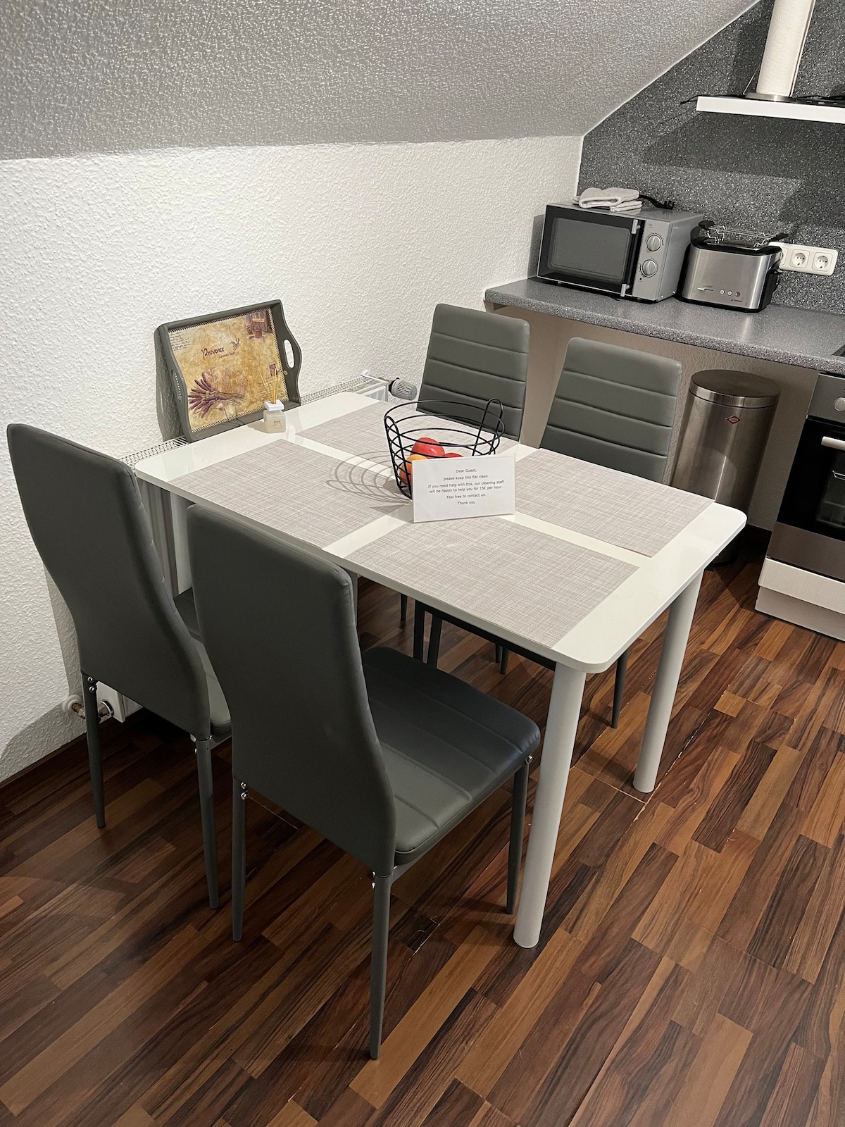 A compact dining area is set with a light-colored table and four grey chairs. A simple decorative basket sits in the center of the table, alongside a menu or information card. The surrounding kitchen appliances and cabinetry are visible in the background.