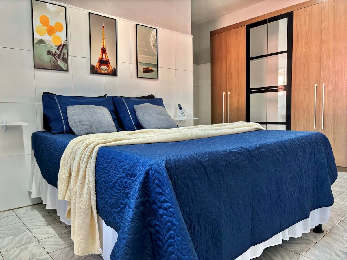 A spacious sleeping area features a large bed dressed in a deep blue quilt and complemented by soft gray pillows. Artwork showcasing iconic landmarks hangs on the wall above the bed. A wardrobe with glass doors is visible to the side.