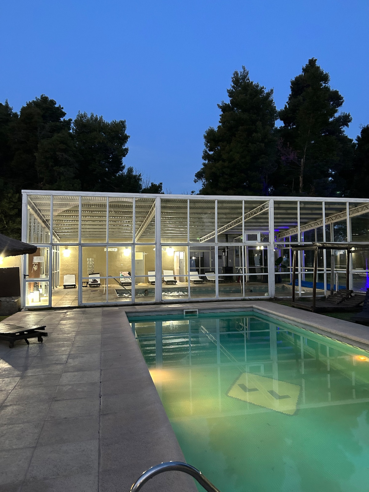 An outdoor pool area is featured, surrounded by tall trees and a glass-enclosed structure housing lounging chairs. The pool's calm water reflects the twilight sky, providing a serene setting for relaxation and leisure.
