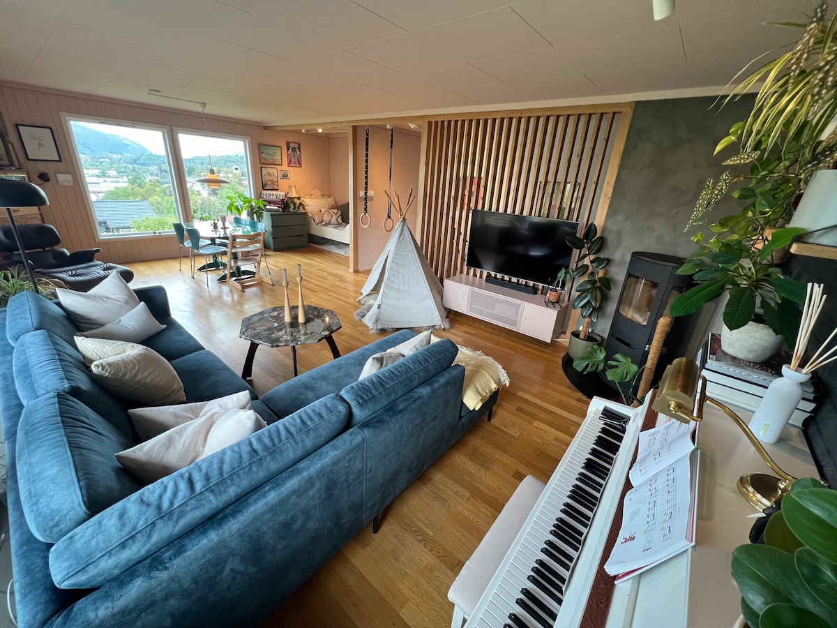 A spacious living room features a blue sectional sofa adorned with cream cushions. A small table is positioned in front, with a piano nearby. Large windows offer views of the landscape, while a television is mounted on a light-colored unit. A stylish play tent adds an element for children.