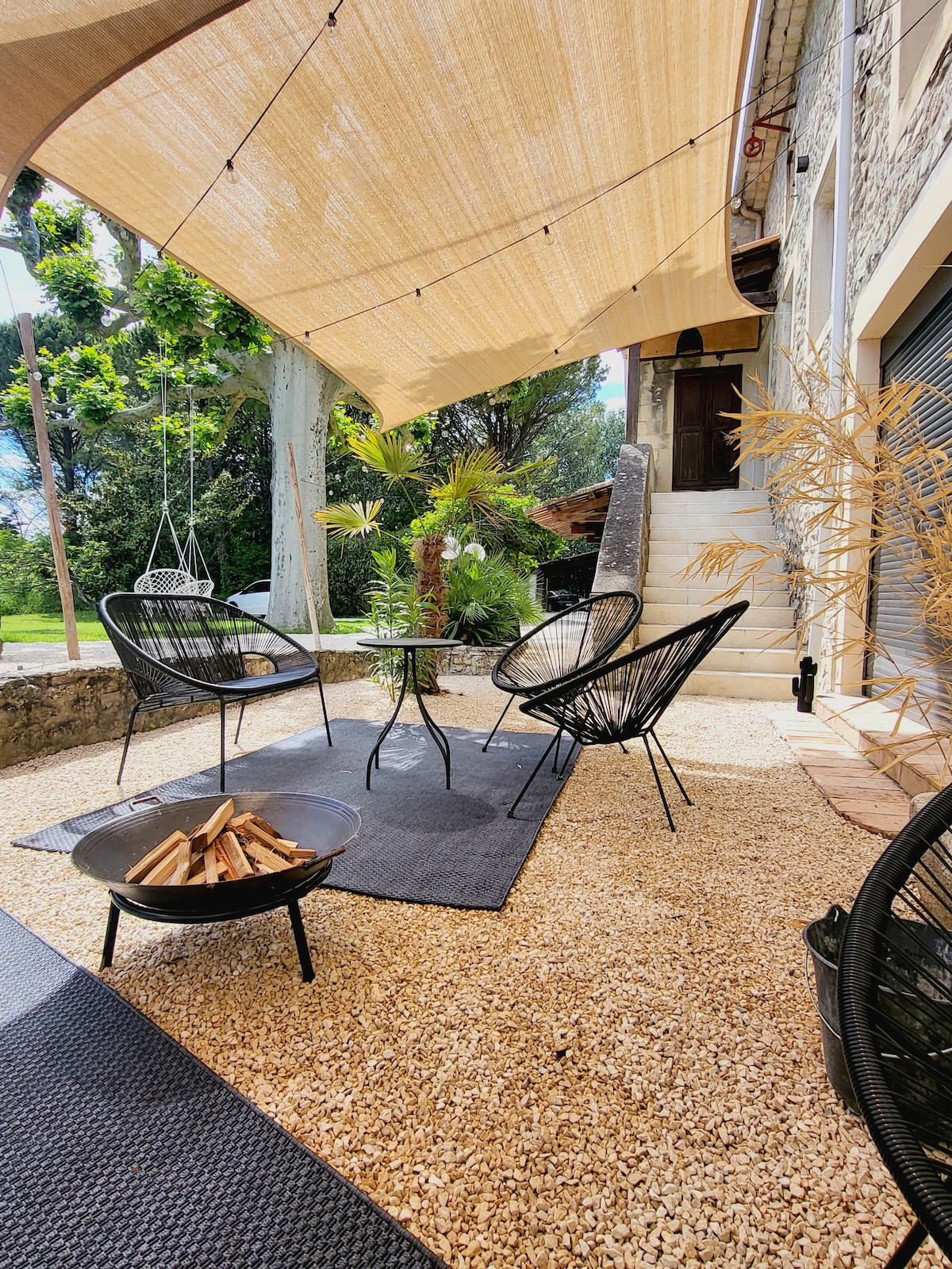 An outdoor seating area features a shaded space with four black chairs surrounding a circular table on a gravel surface. A designated fire pit holds neatly stacked firewood. Decorative plants add a touch of greenery, with steps leading to the entrance of the building.
