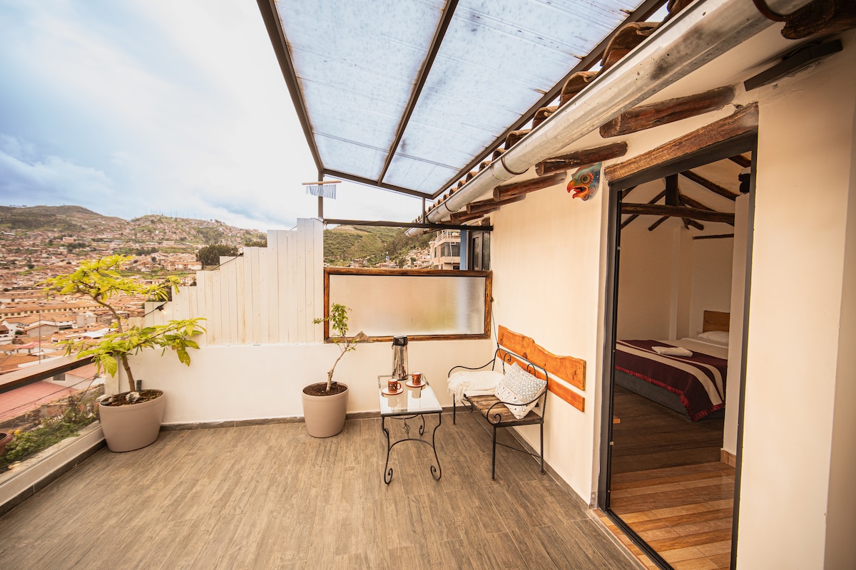 An outdoor terrace is presented with a small table and chair set, surrounded by potted plants. A large window provides views of the Cusco cityscape, while a door on the right leads to an interior space featuring a spacious bed.