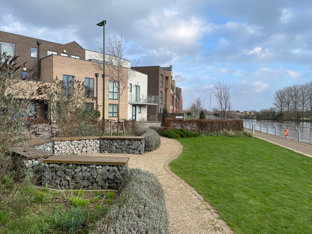 A landscaped outdoor area features a grassy expanse bordered by shrubs and flowering plants. Pathways weave through stone planters, leading toward a riverside view. Modern apartment buildings are visible in the background, set against a cloudy sky and a serene riverbank.