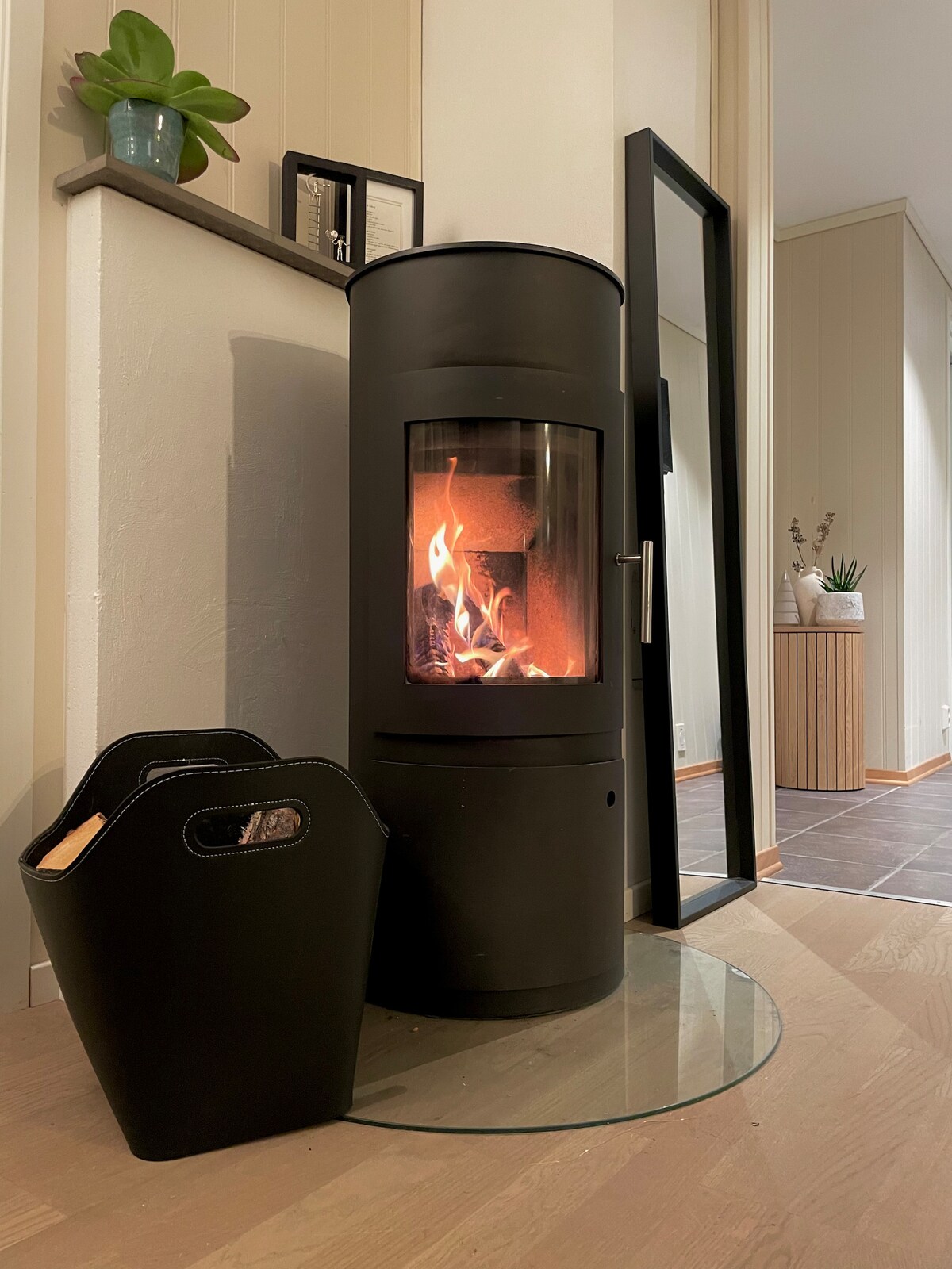 A sleek black stove is positioned against a neutral wall, its glass front revealing a warm, flickering fire. A transparent glass platform protects the floor. Beside the stove, minimalist baskets for firewood are placed for convenience.