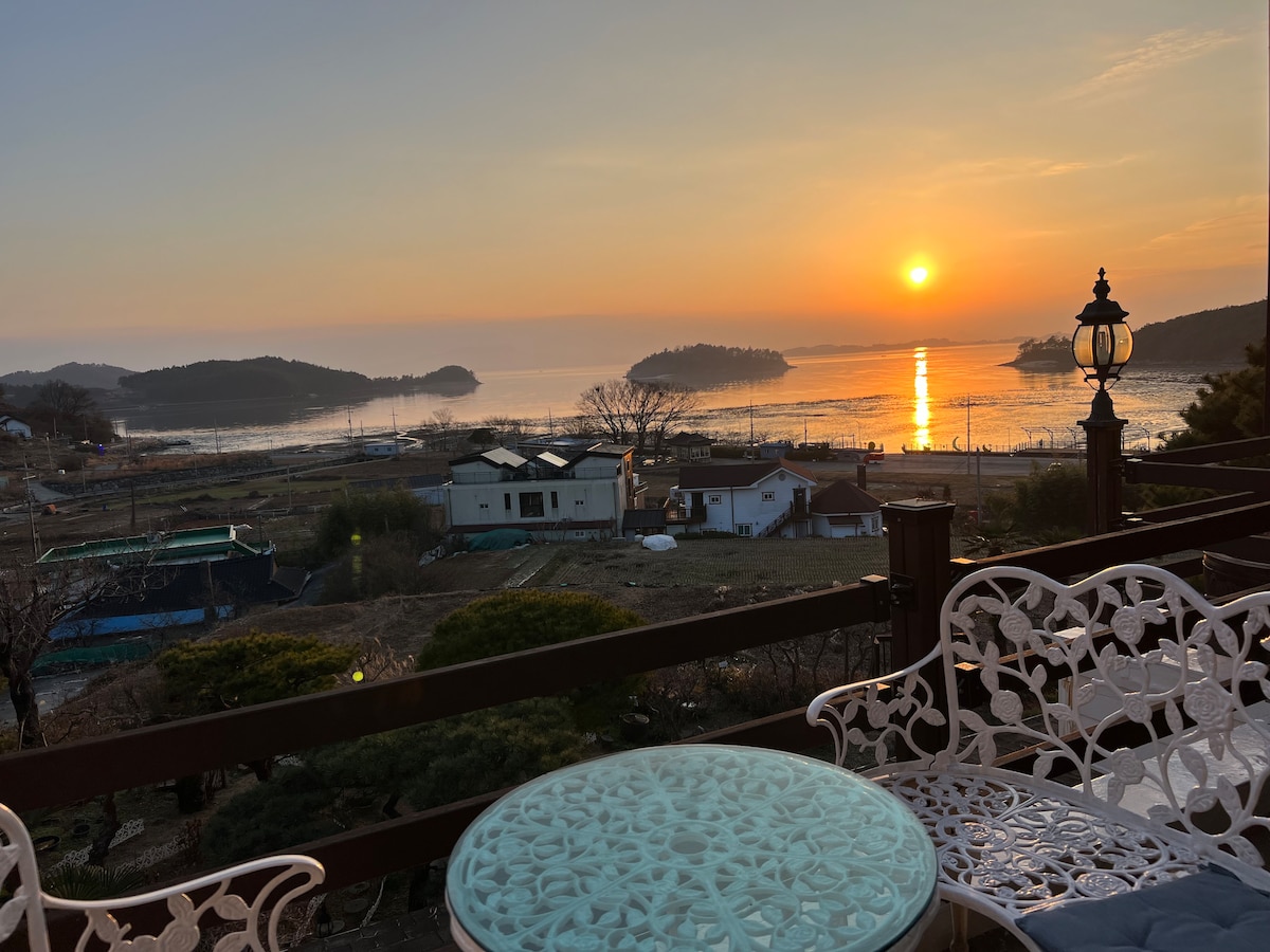 A serene view is captured from a balcony, showcasing a vibrant sunset over calm waters. Two intricately designed white chairs flank a light blue table, inviting relaxation. The landscape includes distant islands and gentle hills, adding depth to the tranquil scene.