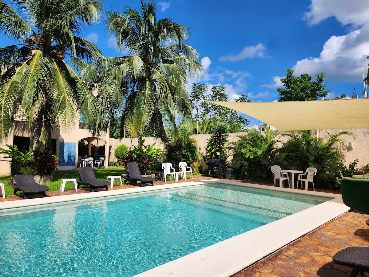 A serene in-ground pool is surrounded by lush greenery and palm trees. Comfortable lounge chairs are positioned beside the pool, with shaded seating nearby. The clear blue sky and bright sun create a refreshing outdoor atmosphere, inviting relaxation and enjoyment in a tropical setting.
