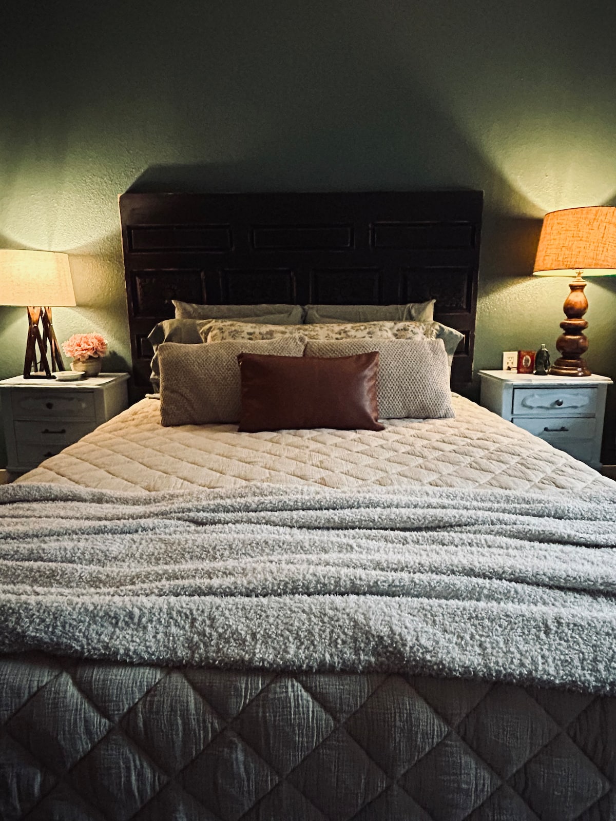 The bedroom features a spacious bed adorned with plush bedding and a decorative throw. Two bedside lamps provide soft lighting on either side, highlighting the dark wood headboard. Neutral tones create a serene atmosphere, complemented by decorative pillows arranged neatly.