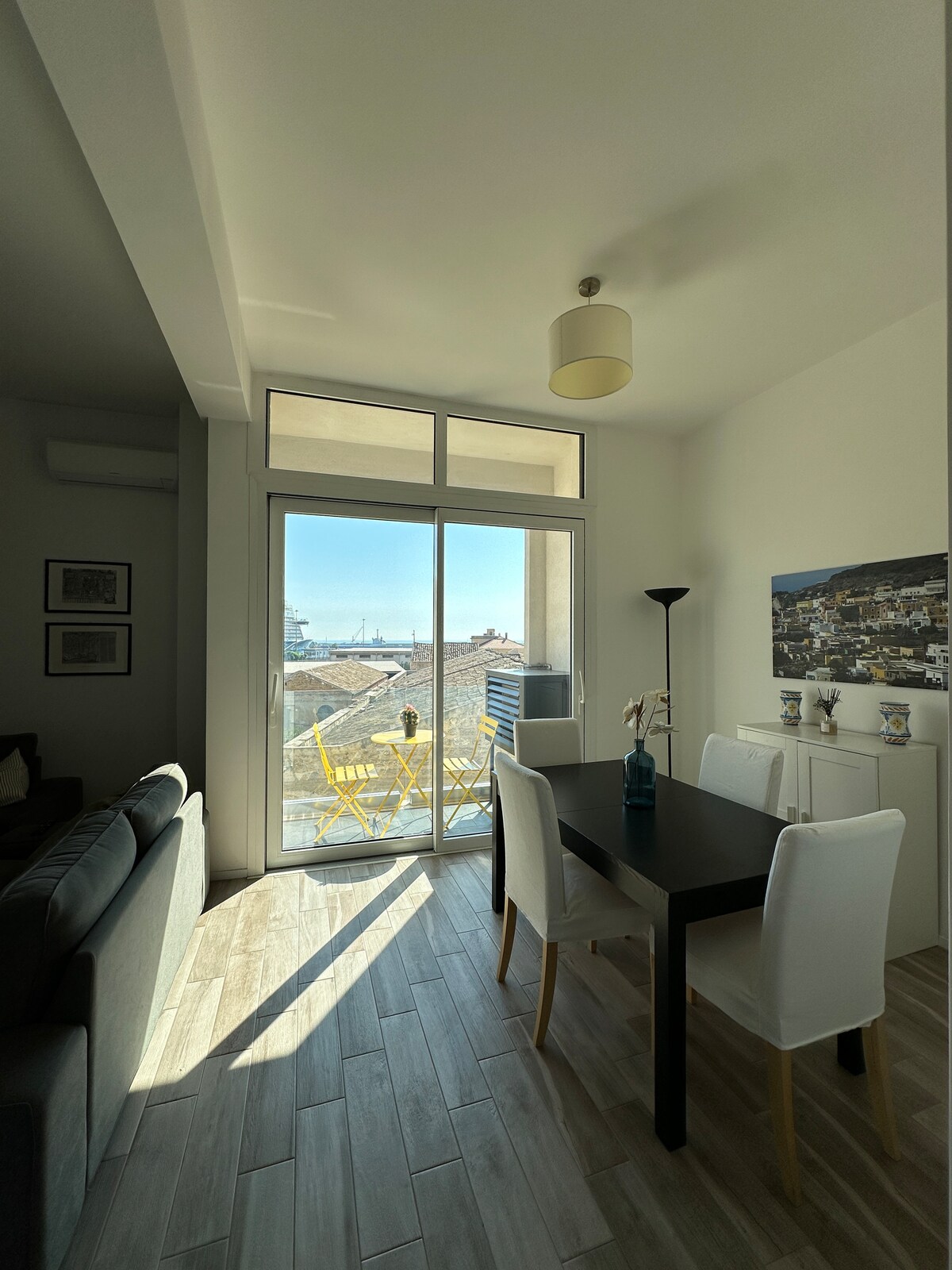 A dining area features a dark table surrounded by white chairs. Natural light floods the space from a large glass door leading to a balcony with two yellow chairs, offering a view of the sea. Light-colored walls and flooring enhance the bright atmosphere.