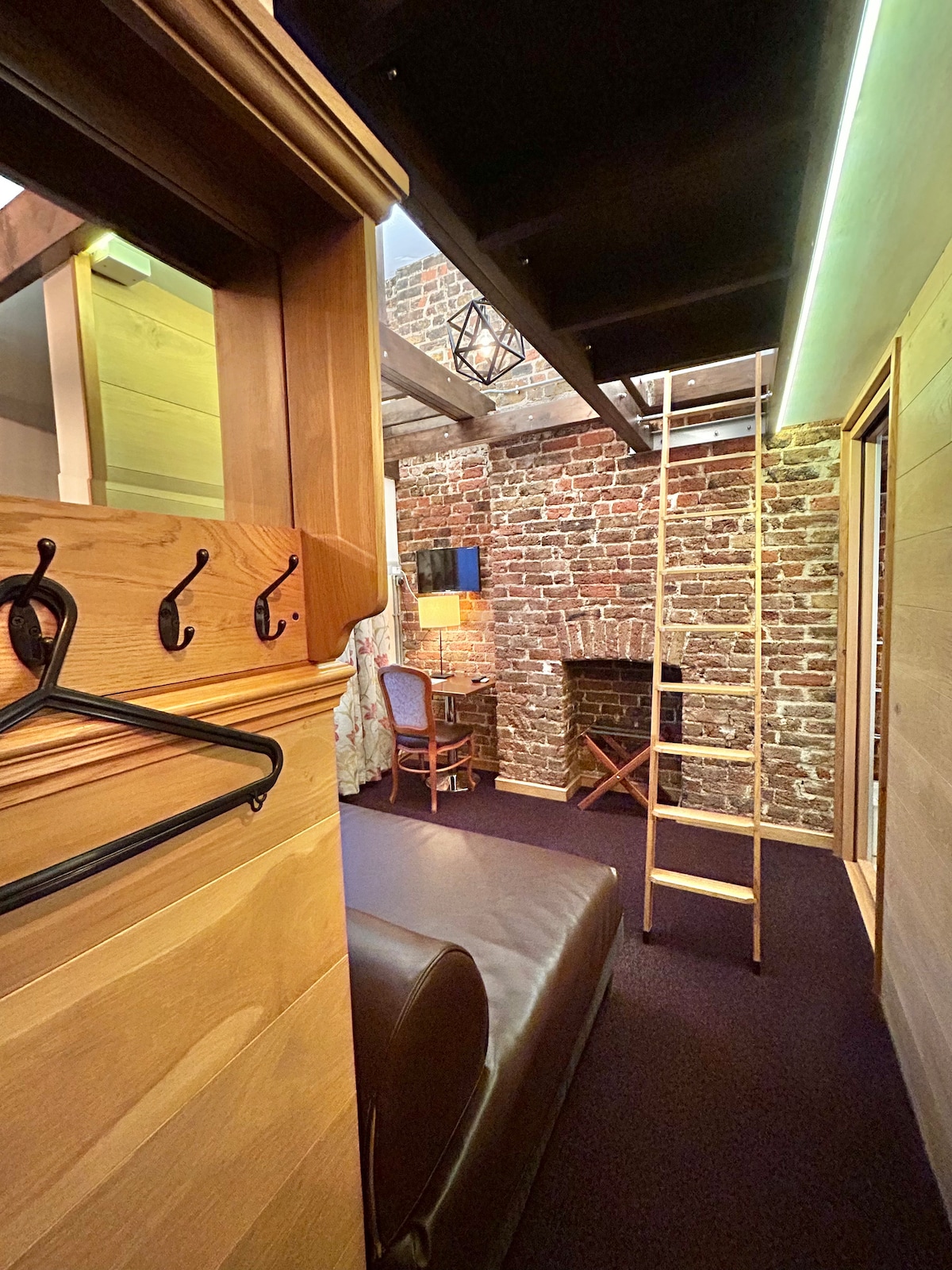 The image captures a view of the loft studio interior, featuring exposed brick walls and natural wood accents. A comfortable sofa is visible, along with a ladder leading up to the mezzanine where the double bed is located. A small lamp and chair are present, enhancing the cozy atmosphere.