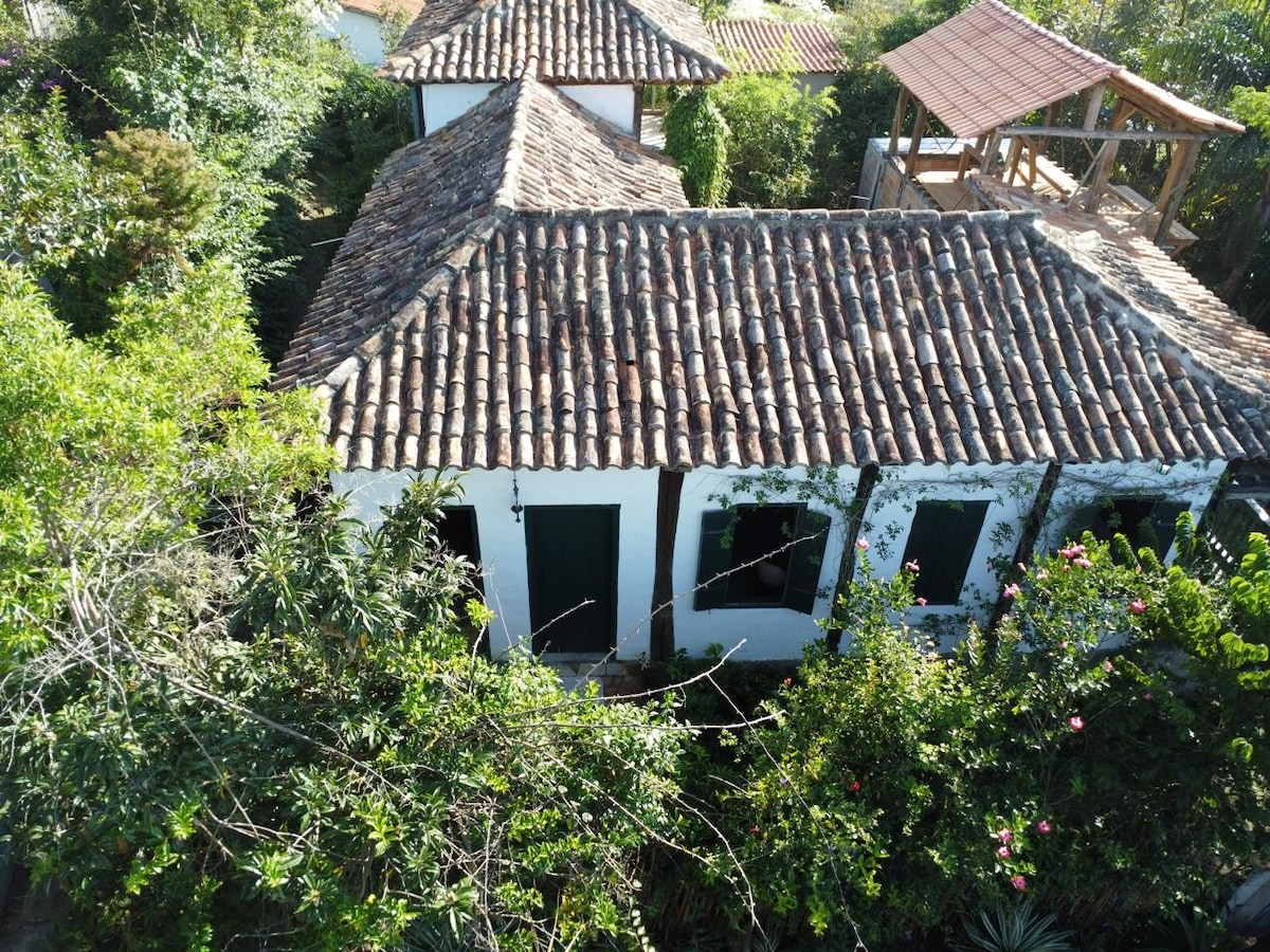 The rustic colonial-style house is framed by lush greenery, showcasing a traditional tiled roof and whitewashed walls. Multiple dark green doors complement the nature surrounding the property, offering glimpses of the tranquil yard and neighboring structures in a peaceful setting.