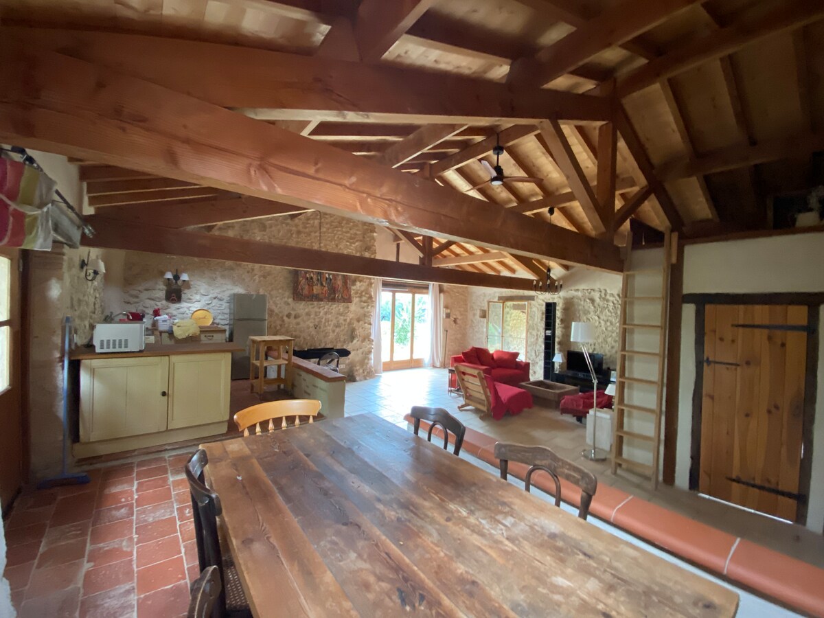 An open living space features a wooden dining table with several chairs, complemented by a cozy seating area with red sofas. Exposed wooden beams create a rustic atmosphere, while large windows offer natural light and views of the garden. The kitchen area is equipped with essential appliances.