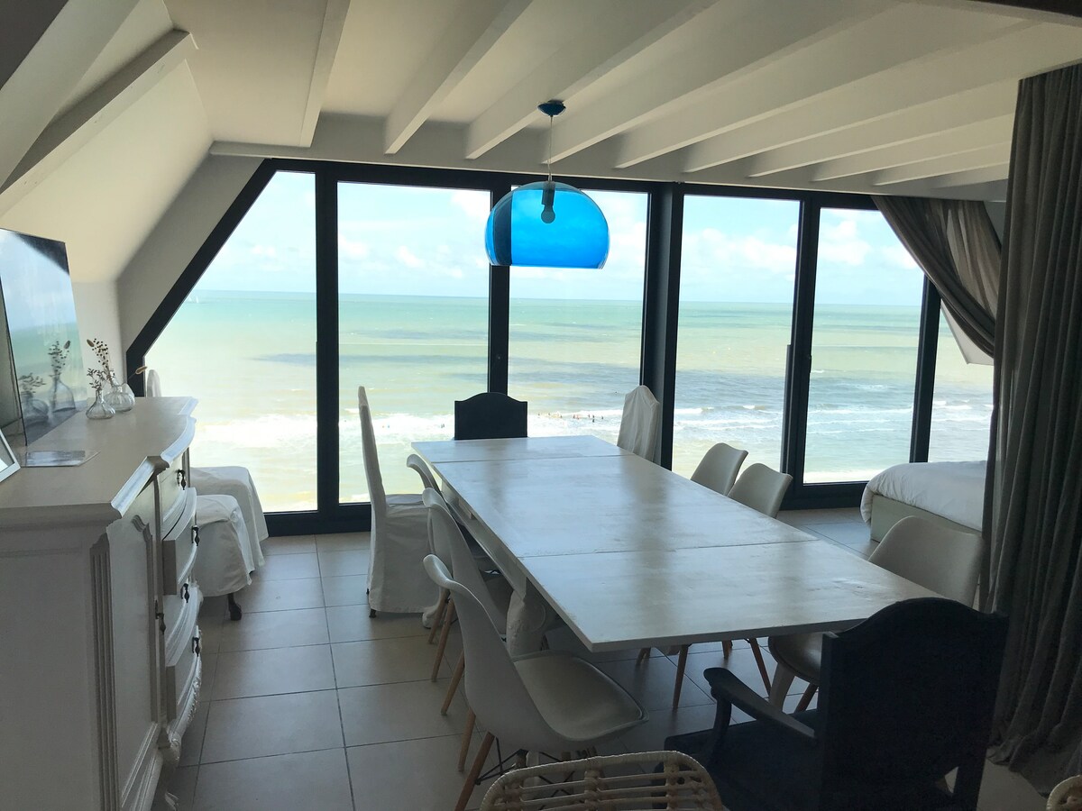 A spacious dining area features a large light-colored table surrounded by white chairs. Floor-to-ceiling windows offer unobstructed views of the sea, with the tranquil blue waters visible and natural light flooding the space. A blue pendant light hangs above the table, enhancing the serene ambiance.