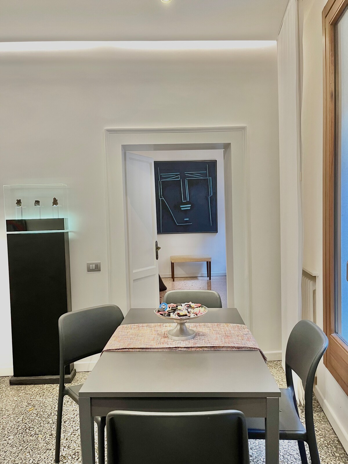 A dining area is showcased with a simple gray table set for meals, complemented by four black chairs. A decorative piece rests on the table, with a glimpse of a doorway leading to another room. A striking wall art piece is visible in the background.